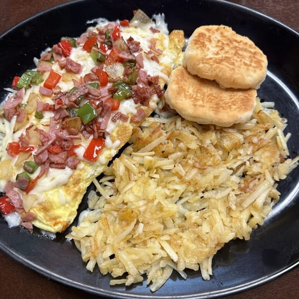 A breakfast plate with a cheesy omelet topped with ham and peppers, hash browns, and biscuits.
